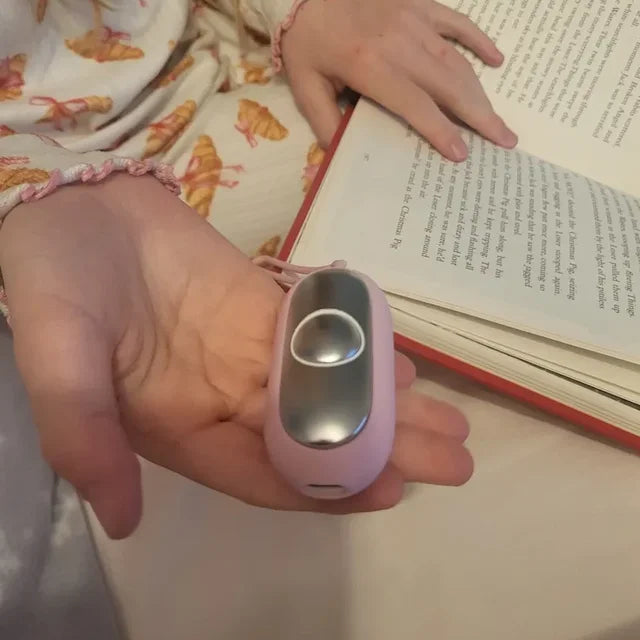 Une enfant tient son CalmKid rose au creux de la main, plaque chromée visible, à côté de son livre.